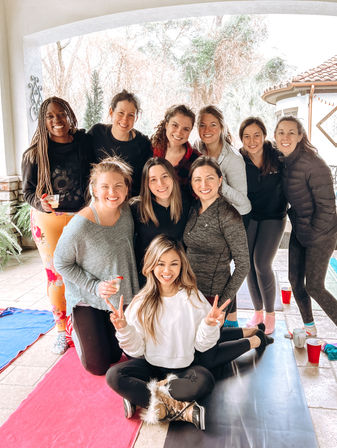 Smiling group of women on a covered backyard patio with yoga mats, drinks, and cozy layers; one woman seated cross‑legged flashing peace signs — outdoor wellness meetup.