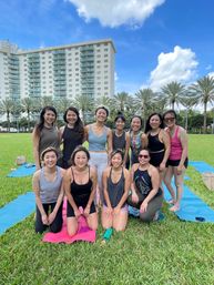 Smiling group of women on yoga mats doing outdoor group yoga in a sunny urban park with palm trees and a high-rise condominium under a blue sky