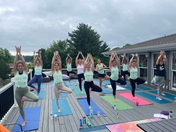 Group of women practicing tree pose on colorful yoga mats during an outdoor rooftop deck yoga class overlooking trees and water under a cloudy sky