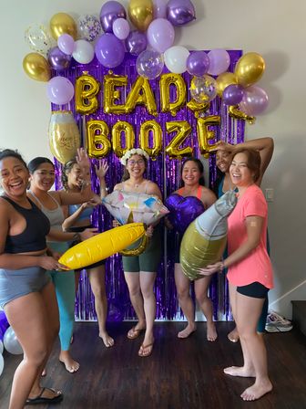 Six smiling women in casual clothes and barefoot pose in front of a purple fringe backdrop with gold letter balloons reading "BEADS BOOZE", surrounded by purple, gold and white balloons while holding oversized inflatable props like a ring and champagne bottle.