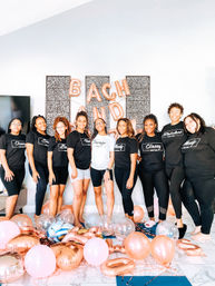 Smiling group of nine women in matching black and white tees pose in a bright modern living room for a bachelorette party surrounded by rose gold and pink balloons and letter balloons