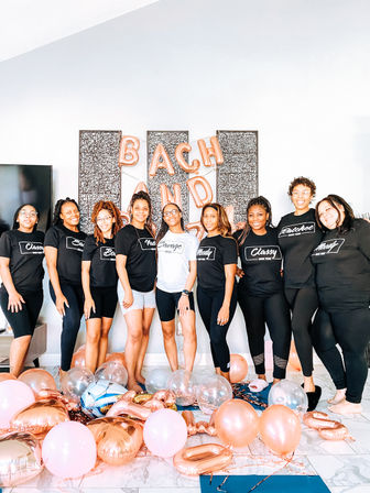 Smiling group of nine women in matching black and white tees pose in a bright modern living room for a bachelorette party surrounded by rose gold and pink balloons and letter balloons