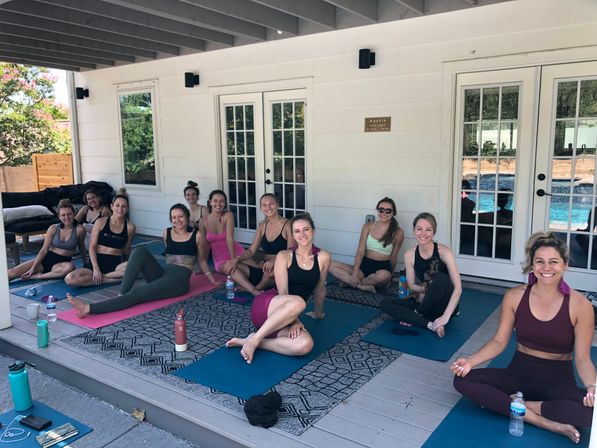 Smiling group of women on yoga mats in a small outdoor yoga class on a covered wooden patio with French doors and a pool visible in the background