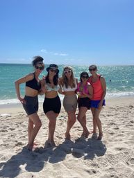 Five friends in swimwear posing with drinks on a sunny sandy beach by the turquoise ocean under a clear blue sky.