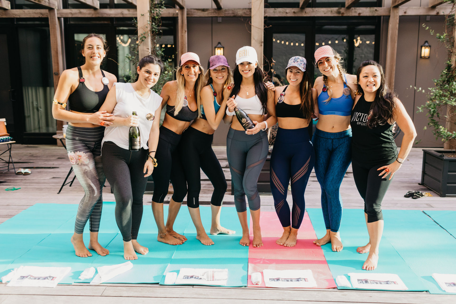 Group of nine women in athletic wear on outdoor wooden deck yoga mats, smiling and holding champagne bottles in a celebratory post-yoga fitness session