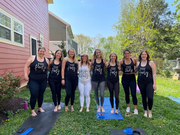 Bachelorette backyard group photo: bride in white and floral crown surrounded by friends in matching black "The Party" tanks, standing on yoga mats on a sunny spring day.
