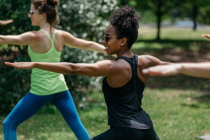 Group of women practicing outdoor yoga in a sunny park, holding Warrior II poses in athletic wear for a group fitness session.