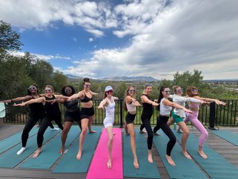 Group of women practicing outdoor yoga in Warrior II on colorful mats on a wooden deck overlooking a valley and distant mountains under a dramatic cloudy sky