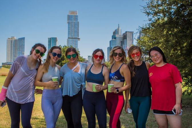 Seven women in colorful activewear and playful sunglasses smiling and holding drinks in a sunny urban park with a modern downtown skyline of high-rise buildings in the background.