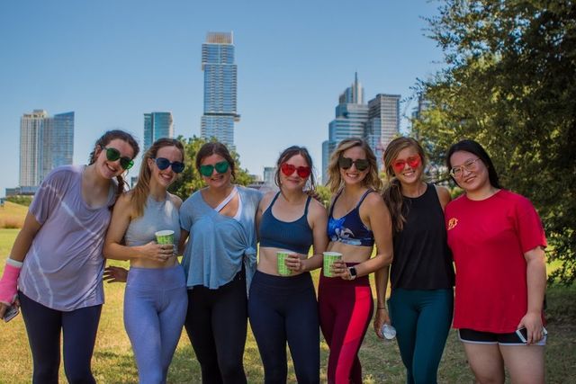 Seven women in colorful activewear and sunglasses smiling and holding drinks while posing in a sunny park with a downtown high-rise skyline (Austin, Texas) in the background.