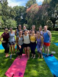 Smiling group of women in activewear posing on colorful yoga mats on a sunny grassy park lawn beneath large trees with pink blooms — outdoor yoga/fitness class