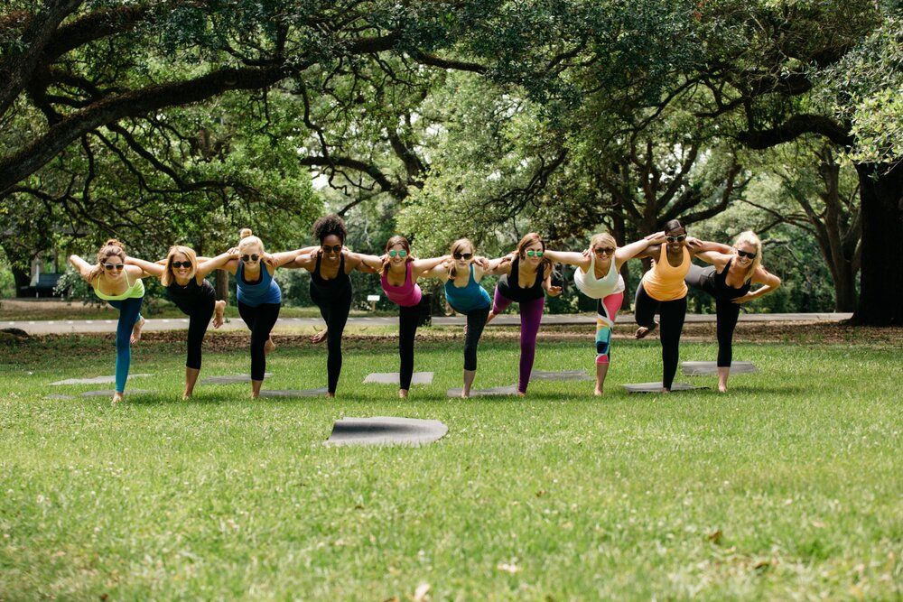 Outdoor yoga class: line of women balancing on one leg, linked arm-in-arm on mats in a grassy park under large shady trees.