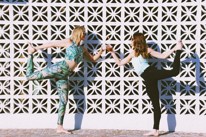 Two women in sunglasses strike mirrored standing-bow yoga poses, clinking drinks while balancing barefoot against a sunlit white patterned breeze-block wall — one in tropical print activewear, the other in black leggings and a light tank.
