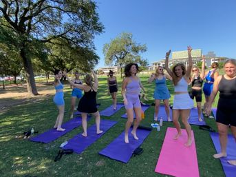 Cheerful outdoor yoga class: group of women in colorful activewear jumping and stretching on purple and pink mats on a sunny park lawn under oak trees with campus buildings in the background.