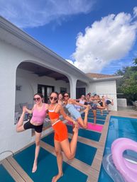 Smiling women practicing poolside outdoor yoga in dancer pose on mats along a backyard patio beside a swimming pool under a bright blue sky.