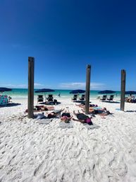 Sun-soaked white-sand beach with turquoise ocean and clear blue sky, weathered wooden posts framing navy umbrellas and chairs, and a group of beachgoers lying on towels sunbathing near the water.