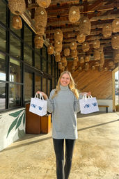 Smiling woman in a gray turtleneck and black leggings holding two white shopping bags with blue logos outside a storefront under a ceiling of woven wicker pendant lights on a sunlit patio.