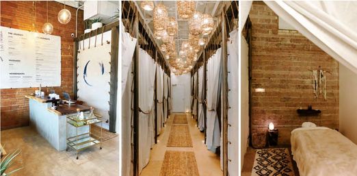 Cozy downtown wellness studio interior with exposed brick, a concrete reception desk, a corridor of curtained massage pods lit by woven rattan pendant lights, and a serene treatment room with a plush massage table and soft ambient lighting.
