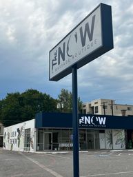 Urban massage boutique storefront with tall roadside sign, deep-blue awning, botanical mural, accessible parking and cloudy sky