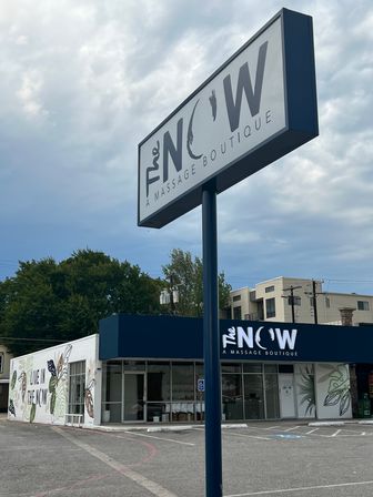 Urban massage boutique storefront with tall roadside sign, deep-blue awning, botanical mural, accessible parking and cloudy sky