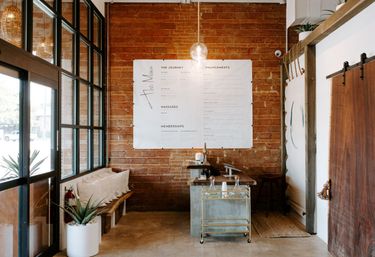 Sunlit industrial-chic spa reception with exposed brick wall, large white menu poster, glass pendant light, bench with pillows, potted succulent by floor-to-ceiling windows and a sliding wooden barn door.
