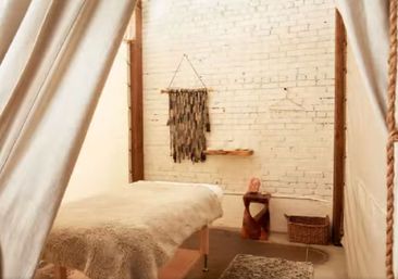 Cozy spa treatment room in an industrial‑chic loft: sheepskin‑covered massage table framed by canvas curtains, white painted brick wall with boho woven wall hanging, wooden shelf, sculptural stool and wicker basket.
