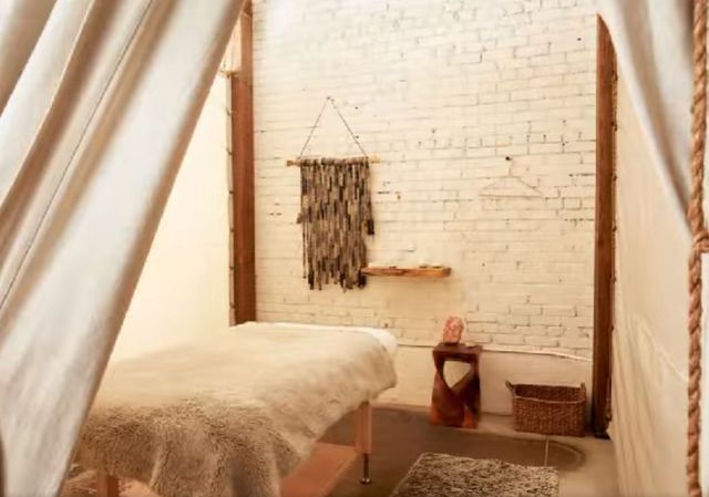 Cozy boho spa treatment room in a loft-style space: canvas tent opening to a massage table draped with a faux-fur throw, white painted brick wall with woven wall hanging, small wooden side table and wicker basket.