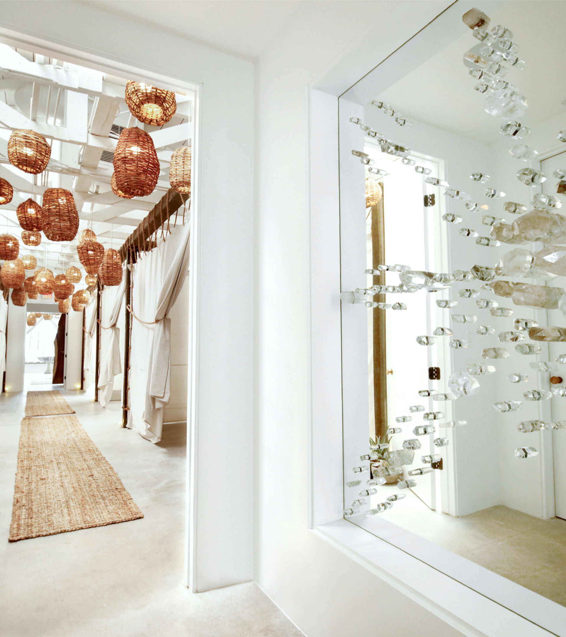 Sunlit coastal boutique spa hallway with clustered woven rattan pendant lights, white curtained treatment cabanas, a jute runner on polished concrete floor, and a crystal wall installation reflected in a mirror.