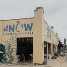 Beige corner storefront advertising a massage boutique with bold blue signage, tropical leaf mural, open entrance with hanging lanterns and potted plants lining the sidewalk.