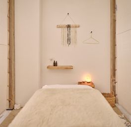 Cozy minimalist spa treatment room with plush white faux-fur massage table, wooden shelf with oil bottles, macramé wall hanging, glowing salt lamp and woven basket