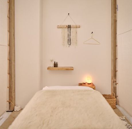 Cozy minimalist spa treatment room with a plush fur-covered massage table, floating wooden shelf with amber oil bottles, glowing salt lamp on a side table, woven wall hanging and neutral boho decor