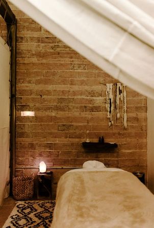 Cozy boho spa treatment room with a plush massage table, exposed brick wall, hanging macramé decor, salt lamp on a wooden side table, woven basket and patterned rug.