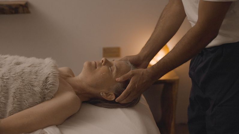 Calm spa treatment: therapist giving a gentle head and temple massage to a relaxed woman on a massage table in a dimly lit room