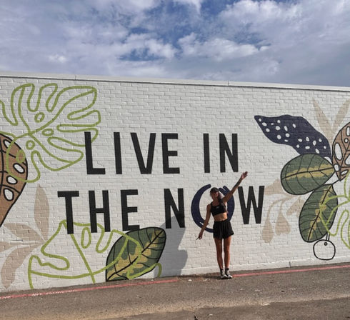 Urban brick wall mural reading LIVE IN THE NOW with oversized tropical leaf illustrations; person in athletic outfit poses on the sidewalk under a partly cloudy blue sky.