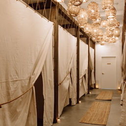 Cozy boho spa hallway with beige linen-curtained changing rooms, hanging woven rattan pendant lights, candles and natural fiber rugs leading to a white door.