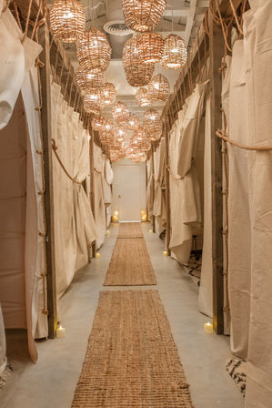Cozy boho wellness hallway lined with curtained changing rooms, glowing rattan pendant lights, jute runners and small candle accents.