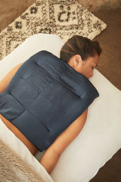 Person lying face down on a white massage table with a navy weighted heat pad across the upper back, cozy rug and faux-fur blanket in a calm at-home spa setting.