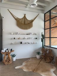Sunlit modern boho retail corner with large macramé wall hanging over white floating shelves of skincare and small goods, sculptural driftwood tables, potted cactus and succulents by a black‑framed window, and a cowhide rug on a concrete floor.