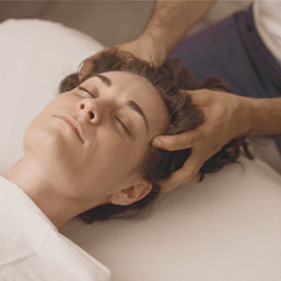 Woman lying with eyes closed while therapist's hands give a soothing scalp massage on a white spa table, relaxation and wellness scene.