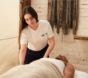 Massage therapist tucking a faux-fur blanket around a relaxed client on a treatment table in a cozy brick‑walled wellness studio