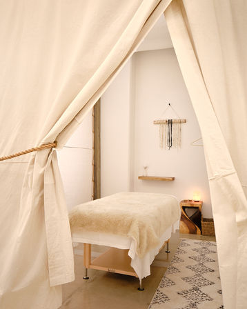 Cozy boho spa treatment tent with a fur-covered massage table, cream canvas curtains tied back, salt lamp on a wooden side table, macramé wall hanging and patterned rug.