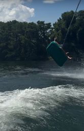 Airborne tow-tube riders launched off a boat wake on a wooded summer lake — green inflatable, splash and blue sky, summer lake tubing and water sports.