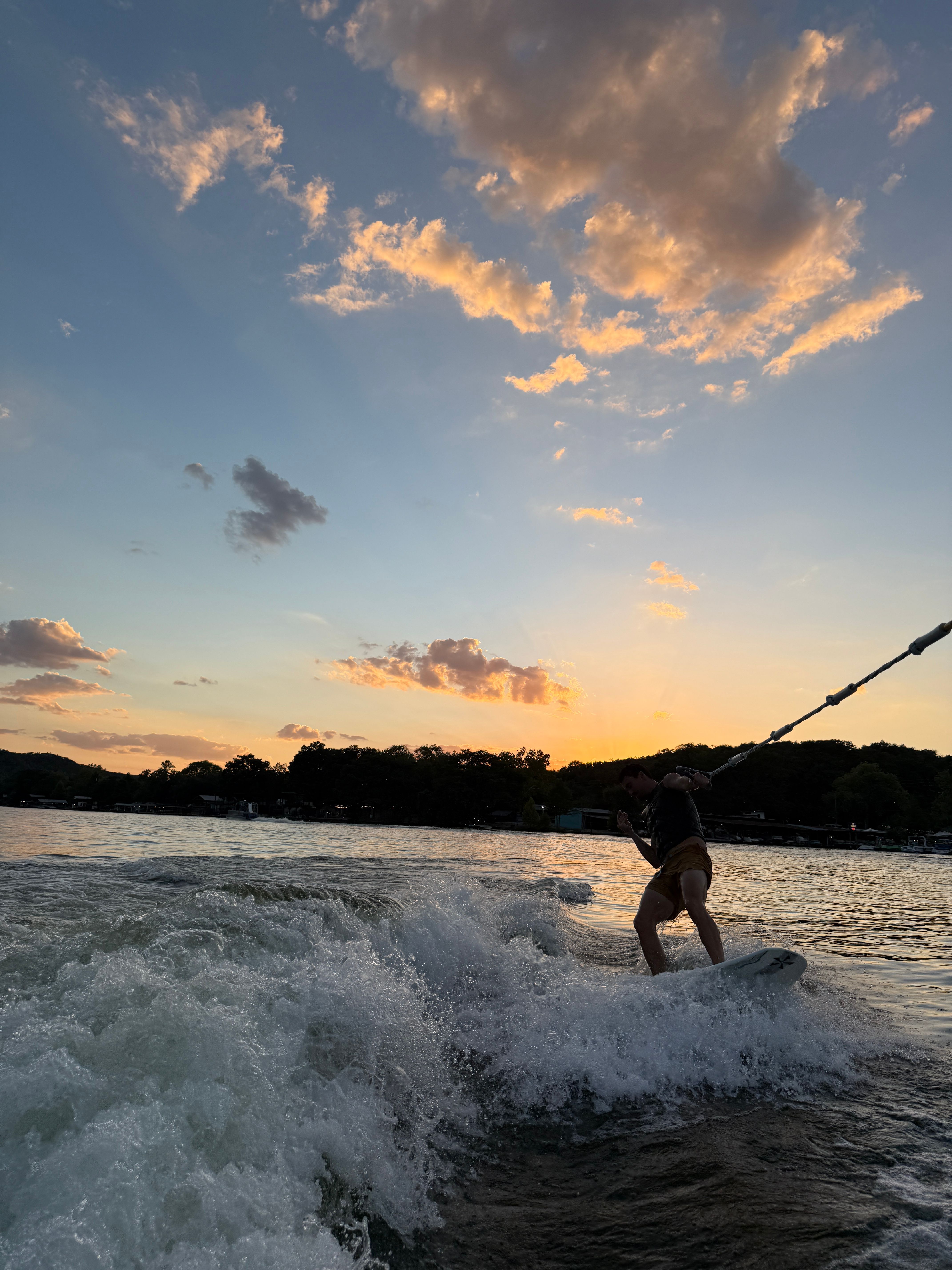 Silhouetted wakeboarder carving a boat wake on a lake at sunset, splashing white water beneath golden clouds and a colorful sky