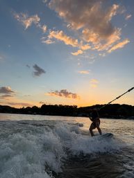 Silhouetted wakeboarder carving a boat wake on a lake at sunset, splashing white water beneath golden clouds and a colorful sky