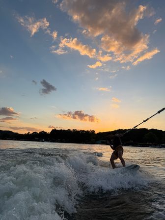 Silhouetted wakeboarder carving a boat wake on a lake at sunset, splashing white water beneath golden clouds and a colorful sky