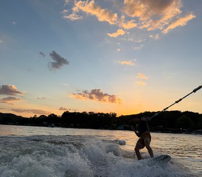 Silhouetted wakeboarder carving a boat wake on a lake at sunset, splashing white water beneath golden clouds and a colorful sky