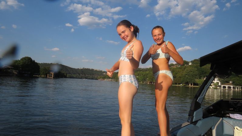 Two smiling girls in swimsuits giving thumbs-up on the edge of a speedboat during a sunny summer lake day, blue sky, tree-covered hills and waterfront homes in the background.