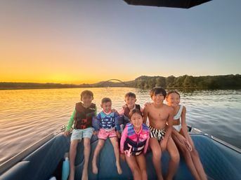 Six kids smiling on a boat at sunset on a calm lake, wearing swimsuits and life jackets with a tree-lined shore and arched bridge on the horizon — golden sky and reflective water.