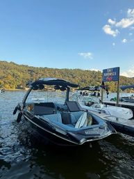 Black wakeboard-style boat moored at a lakeside dock with other boats nearby, tree-covered hills and clear blue sky