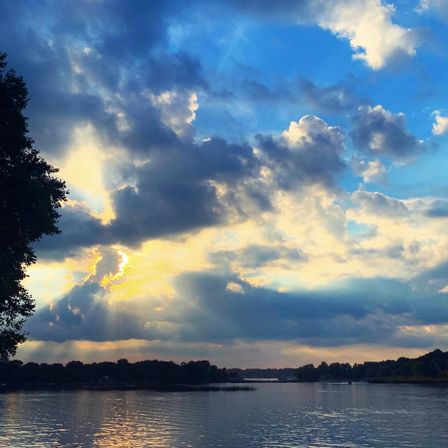 Sunset sky with dramatic cumulus clouds and golden sunbeams over a calm lake, treeline silhouette reflected in rippling water.
